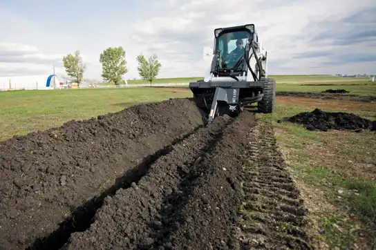Compact track loader with a trencher attachment, actively digging a deep, narrow trench in a field for utility lines in Roseburg, Oregon.